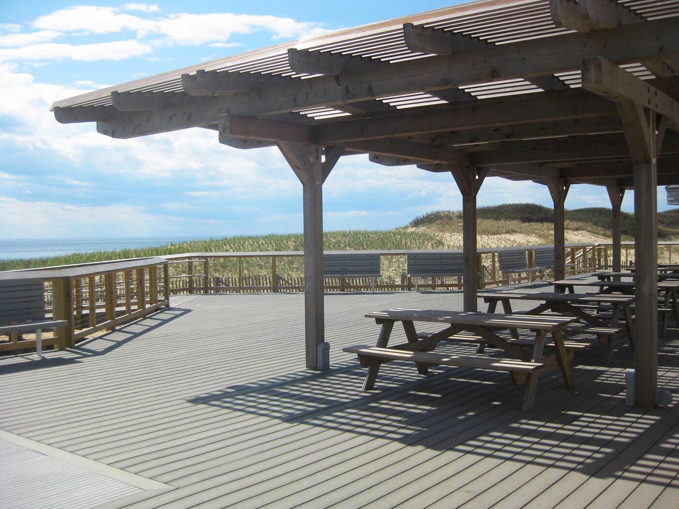 A wooden pergola with picnic tables on a deck overlooking the ocean.