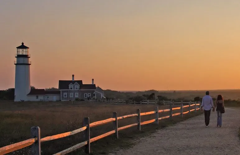 A couple walks along a pathway towards a lighthouse at sunset.
