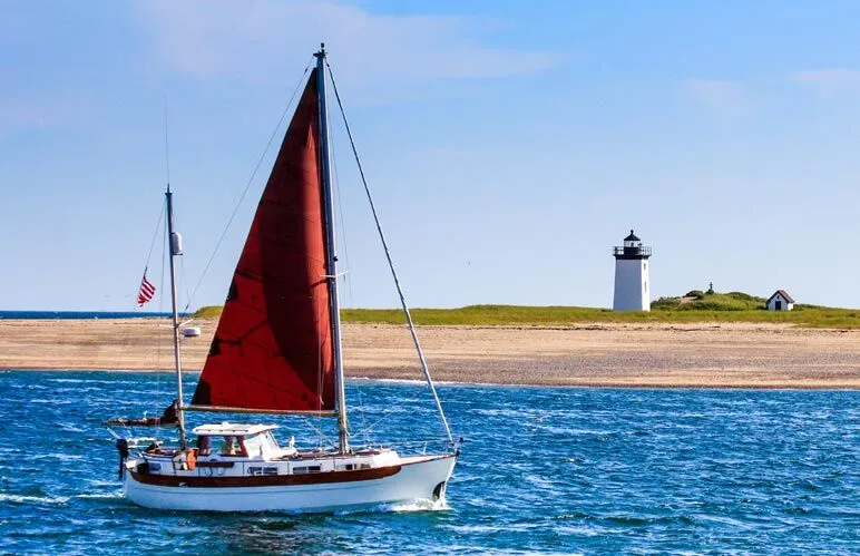 A sailboat with a red sail in the ocean near a beach with a lighthouse.